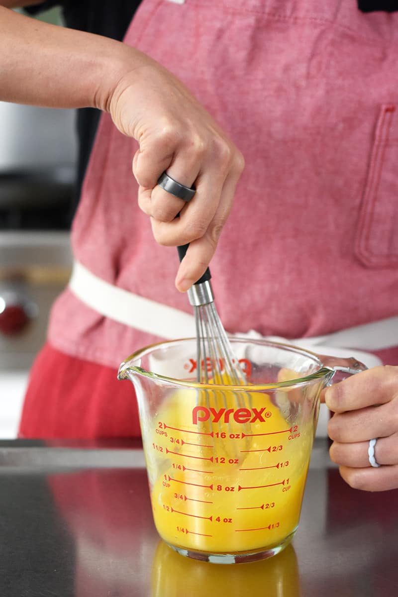 A person in a red apron is whisking eggs and salt in a 2-cup Pyrex glass measuring cup.