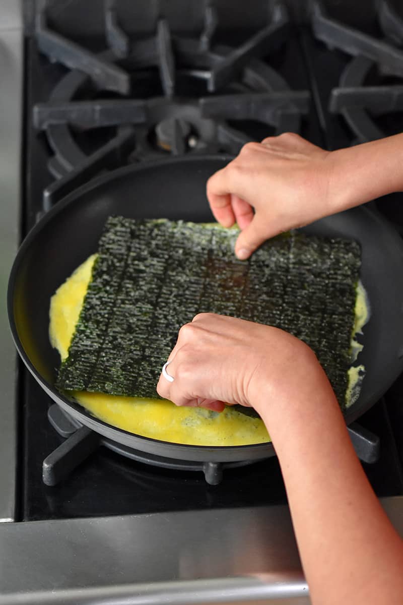 Two hands placing a rectangular piece of roasted seaweed on a thin layer of cooked egg in a non-stick skillet.