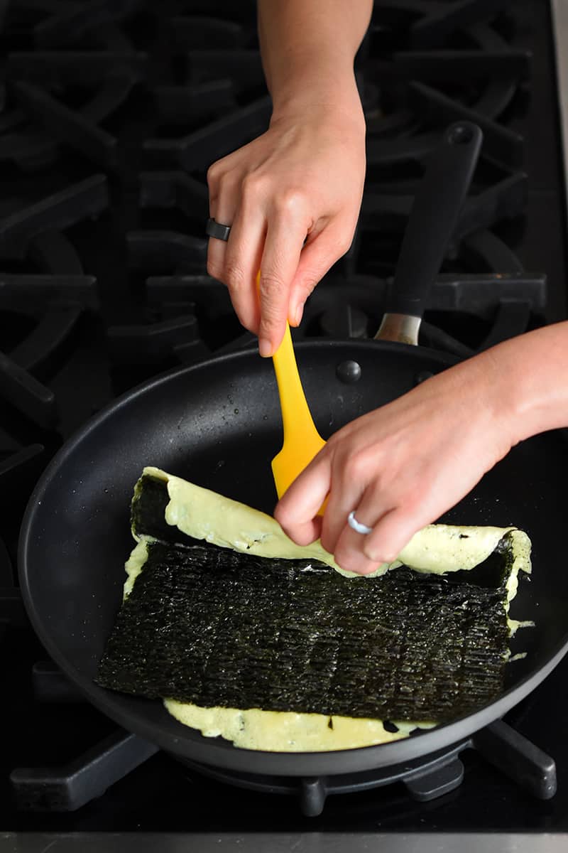 Two hands are using a yellow silicone spatula to fold over the edge of an egg omelet topped with seaweed.