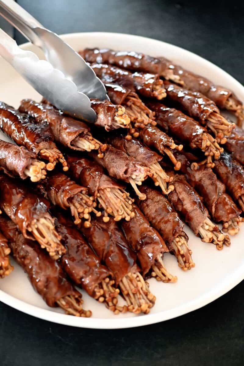 A white plate filled with a large stack of cooked beef and enoki mushroom rolls