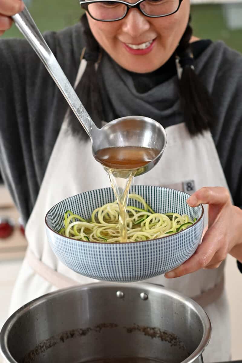 A smiling Asian woman is ladling hot beef broth onto a bowl of spiralized zucchini noodles