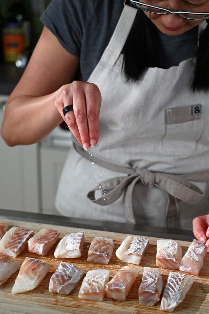 Sprinkling salt on a wooden cutting board with cod fish sticks on it.