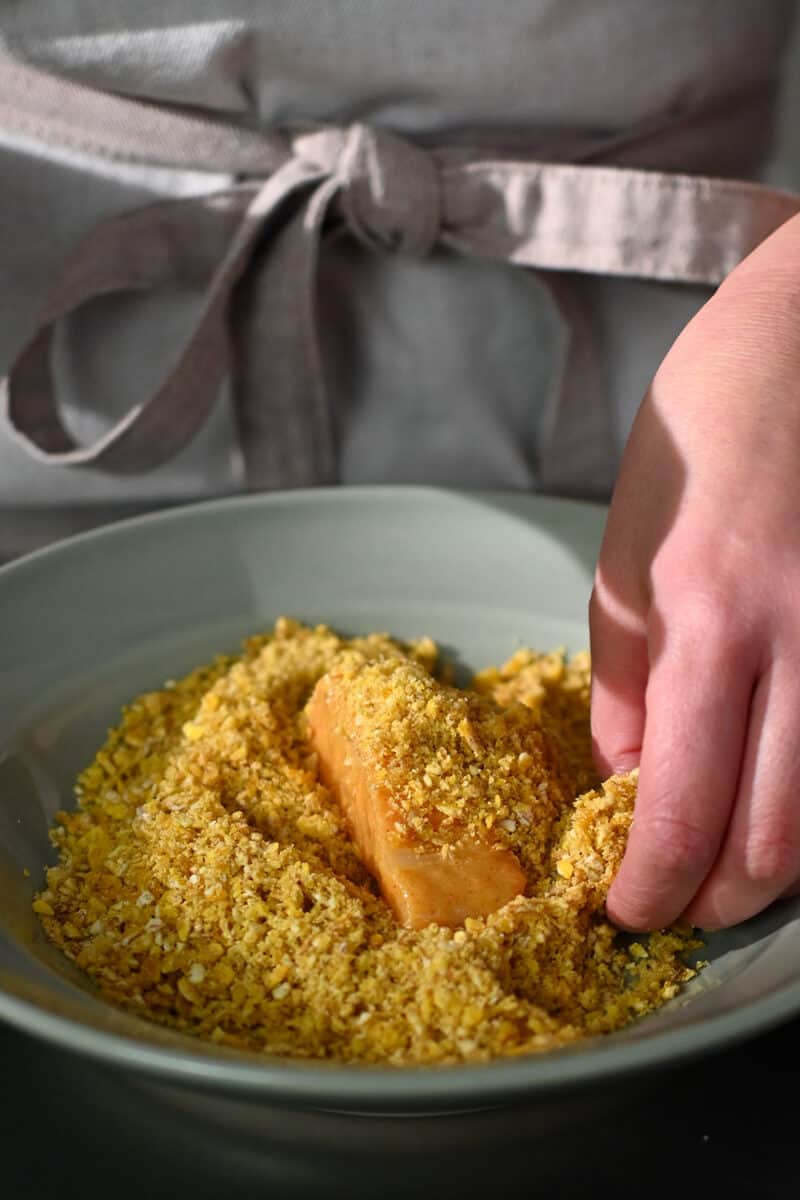A fish fillet is in a shallow dish with plantain chip crumbs.