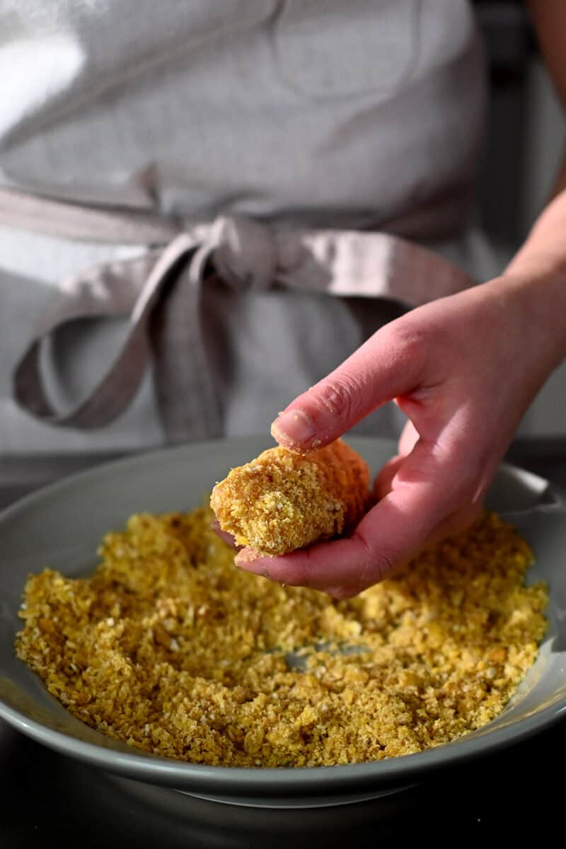 A hand is coating a fish stick with plantain chip crumbs