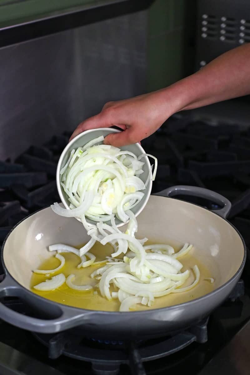 Adding a small bowl of sliced onions to a pan filled with olive oil.