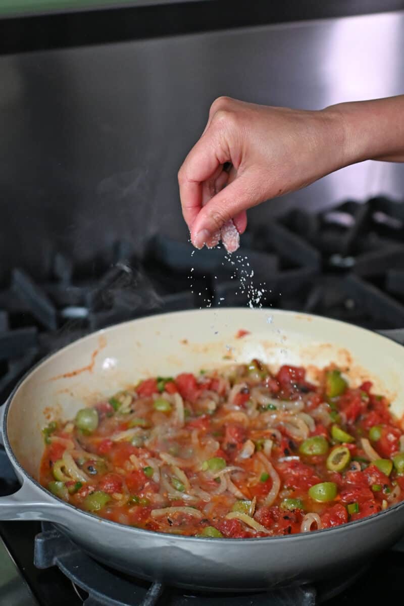 Adding a sprinkle of salt to Veracruz sauce in a skillet.