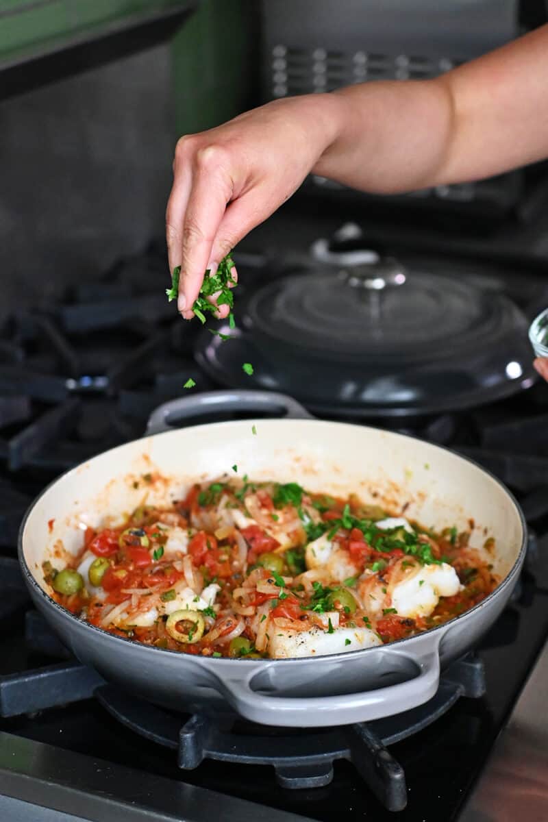 A hand is sprinkling minced fresh cilantro on a pan filled with white fish in Veracruz sauce