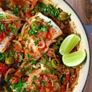 An overhead shot of white fish Veracruz in a skillet with sliced lime wedges on the side.