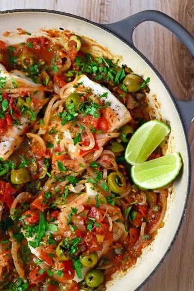 An overhead shot of white fish Veracruz in a skillet with sliced lime wedges on the side.