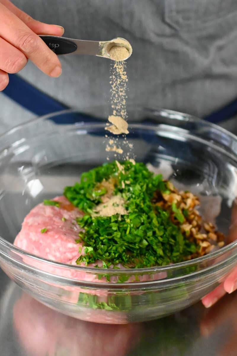 Adding a spoonful of white pepper to a bowl filled with ingredients for wonton meatballs.