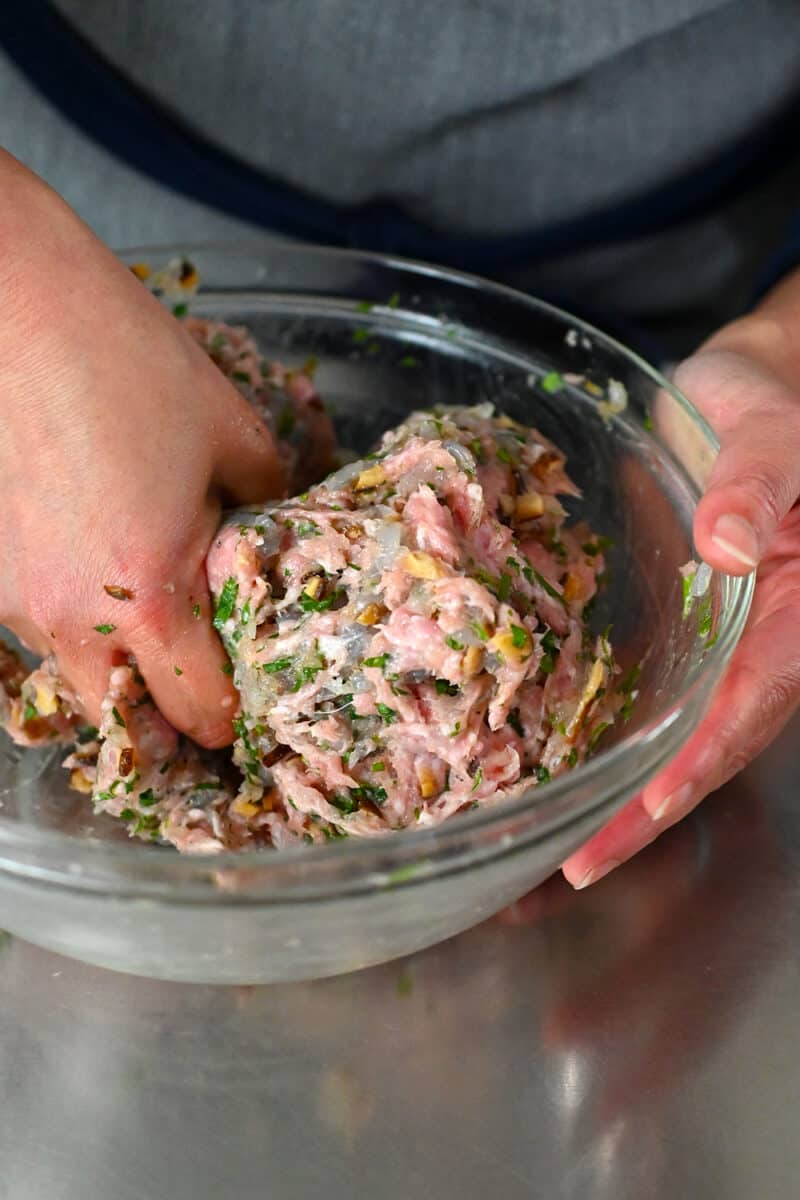 A hand is mixing and squeezing the wonton meatball ingredients in a large glass mixing bowl.