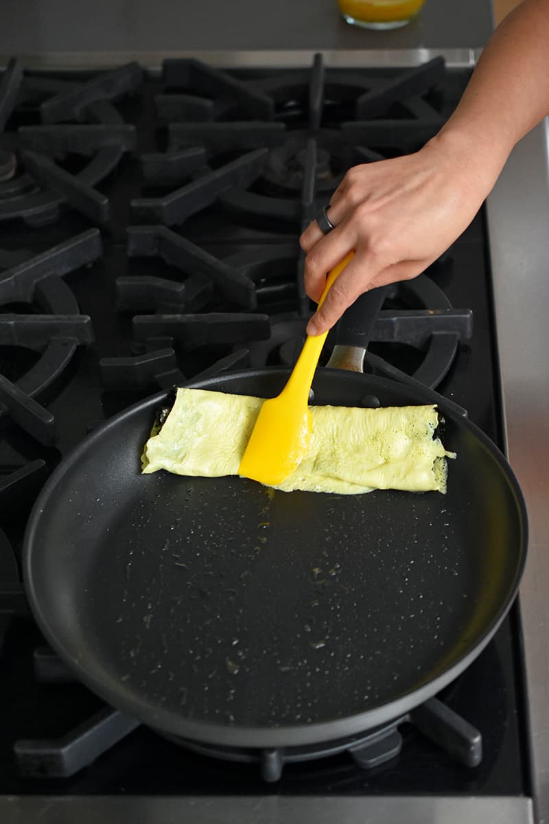 A hand holding a yellow silicone spatula is pushing a rolled egg omelet with seaweed to the edge of a large non-stick skillet.