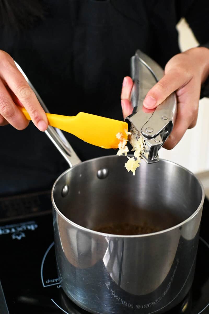 Adding minced garlic to a small saucepan with the ingredients for honey sriracha glaze.