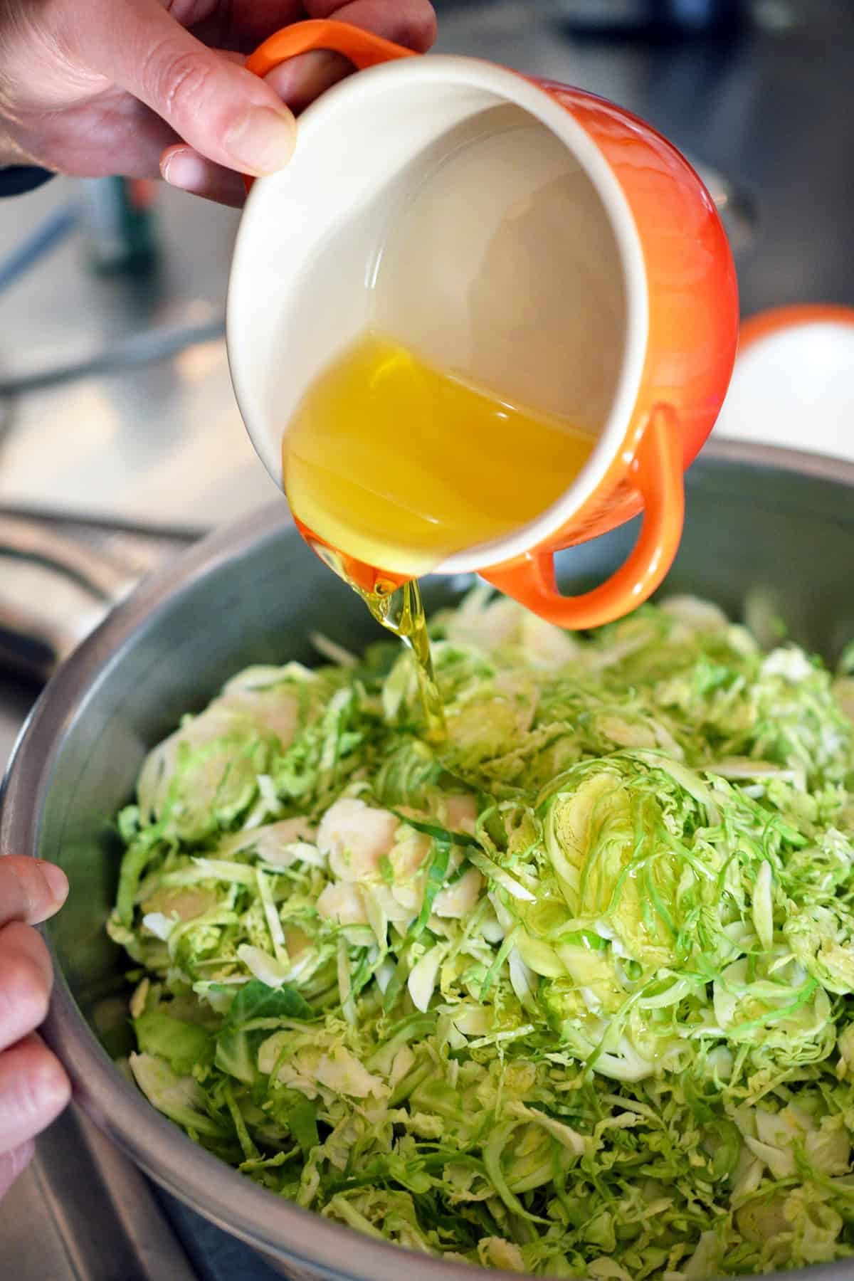 Pouring melted ghee from a small orange ramekin into a bowl of shredded raw Brussels sprouts.