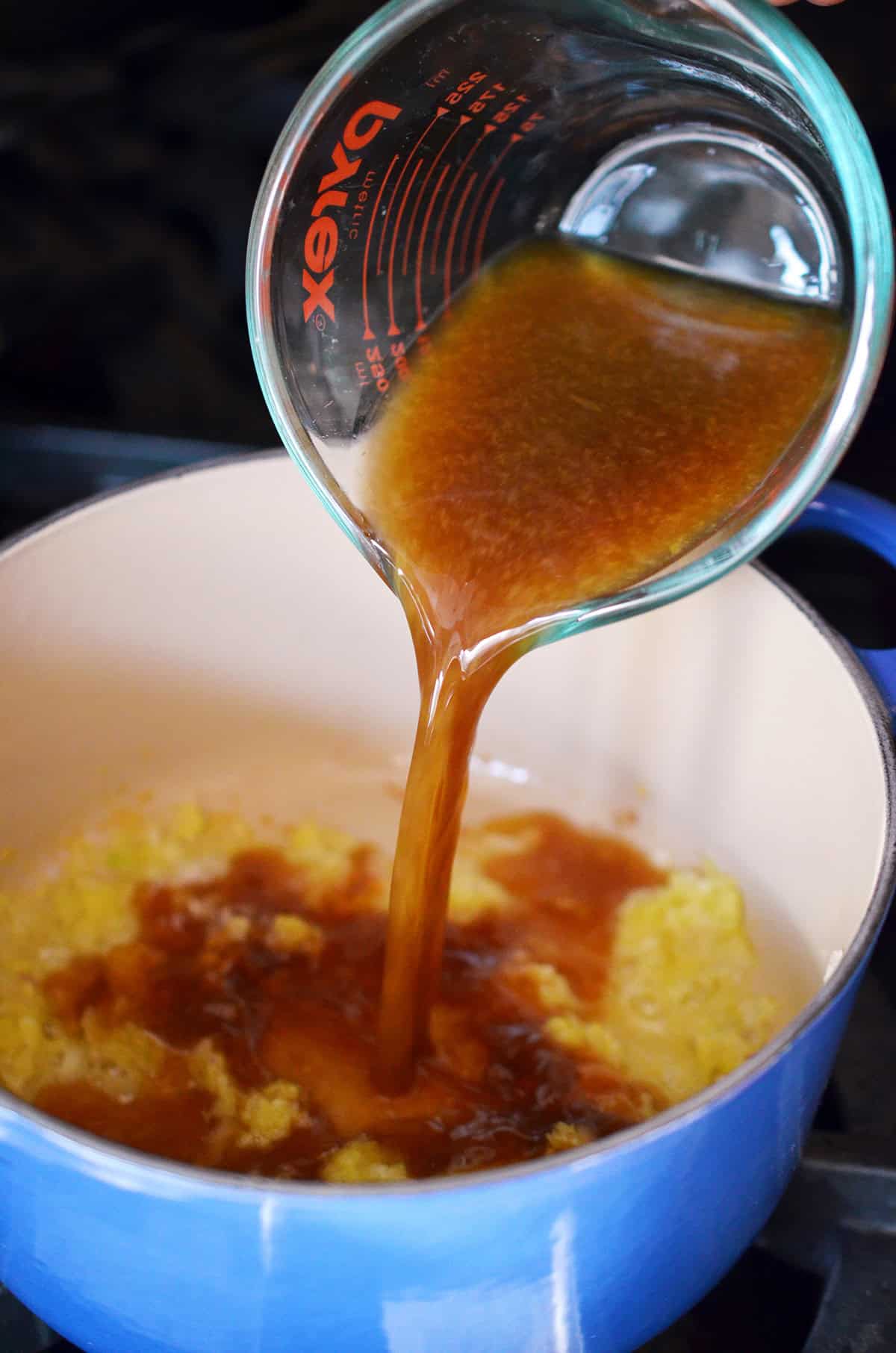 Pouring a brown liquid into a blue saucepan with sautéed ginger, garlic, and shallots.
