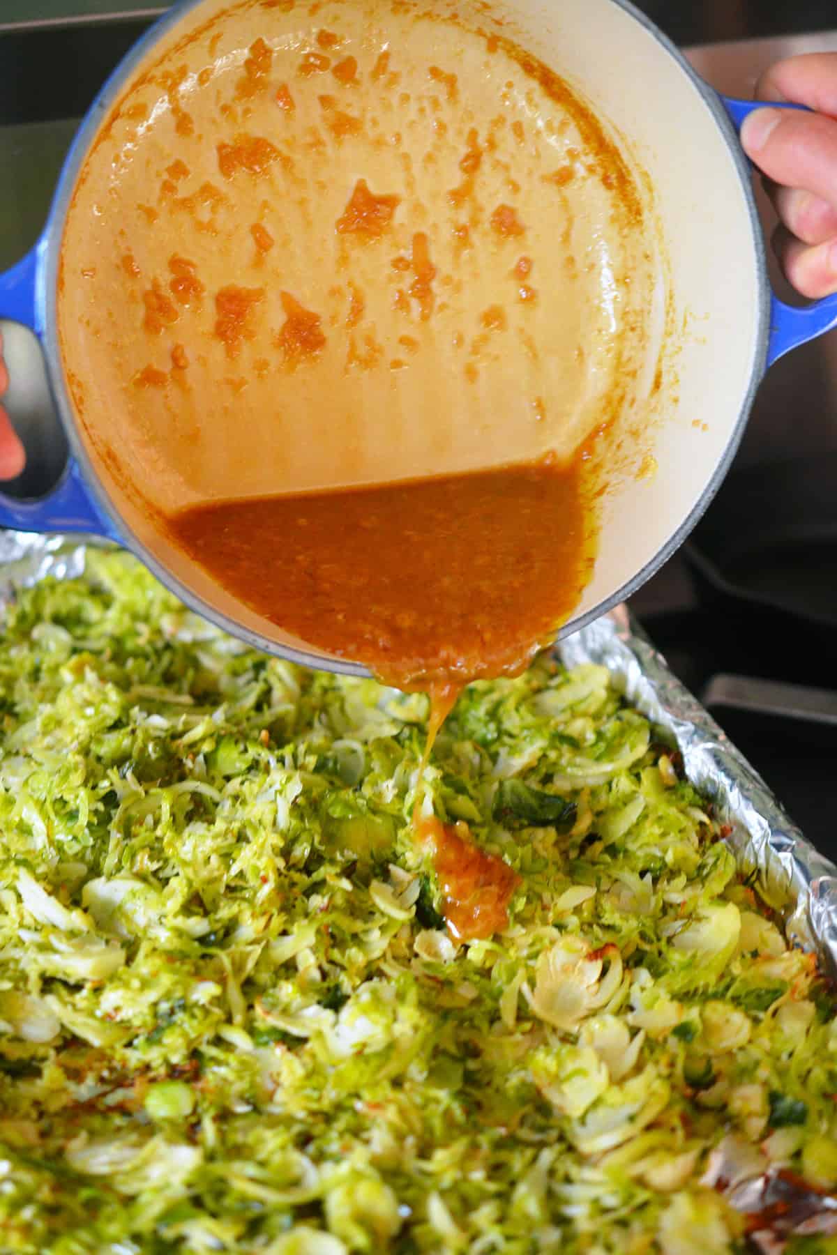Pouring an Asian citrus dressing from a blue saucepan onto a rimmed baking sheet of roasted shaved Brussels sprouts.