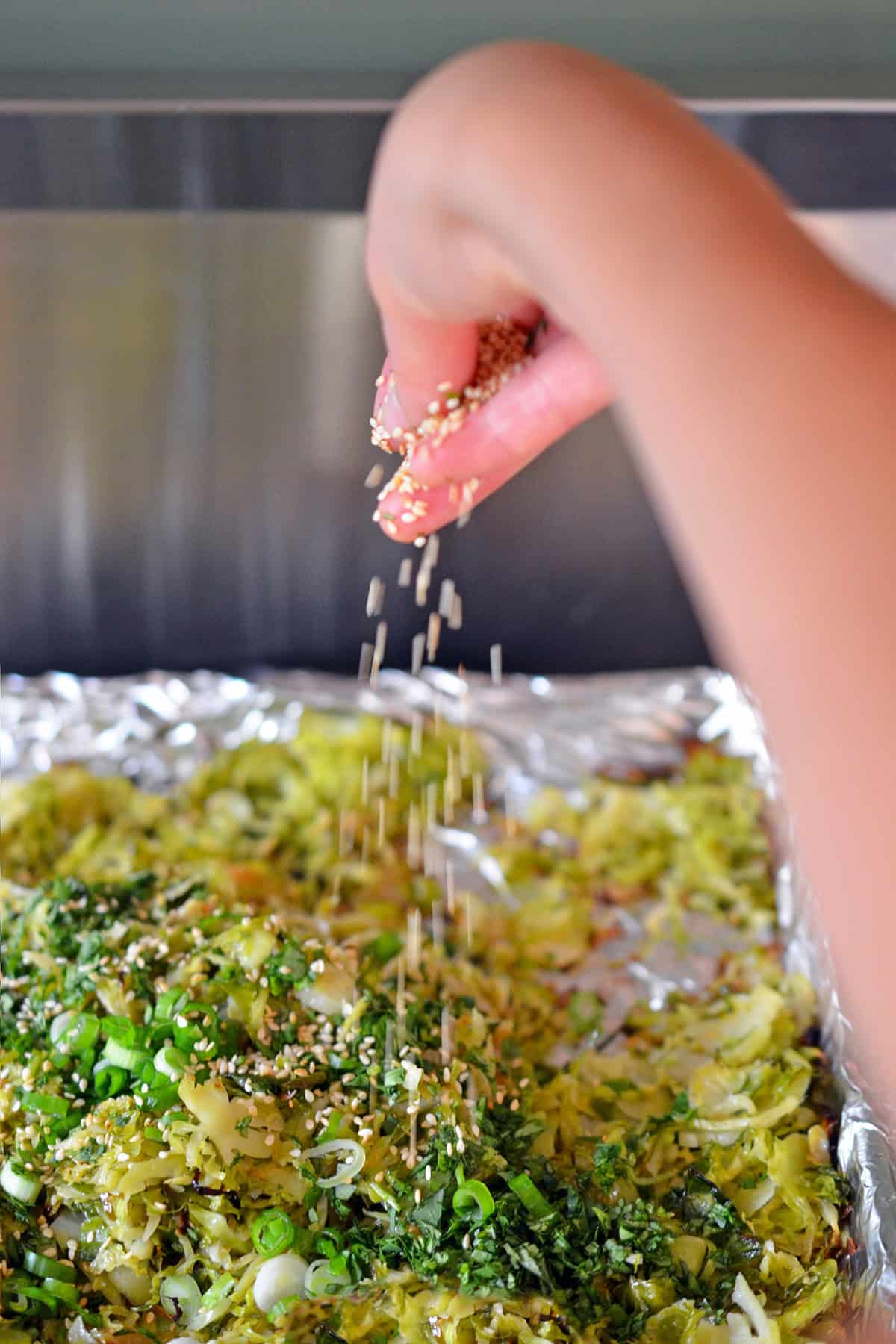 A hand is sprinkling toasted sesame seeds on a rimmed baking sheet with roasted shaved Brussels sprouts and fresh herbs.