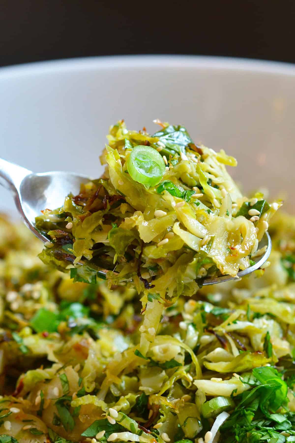 A closeup of a spoon lifting up a serving of roasted Brussels sprouts salad with Asian dressing from a white bowl.