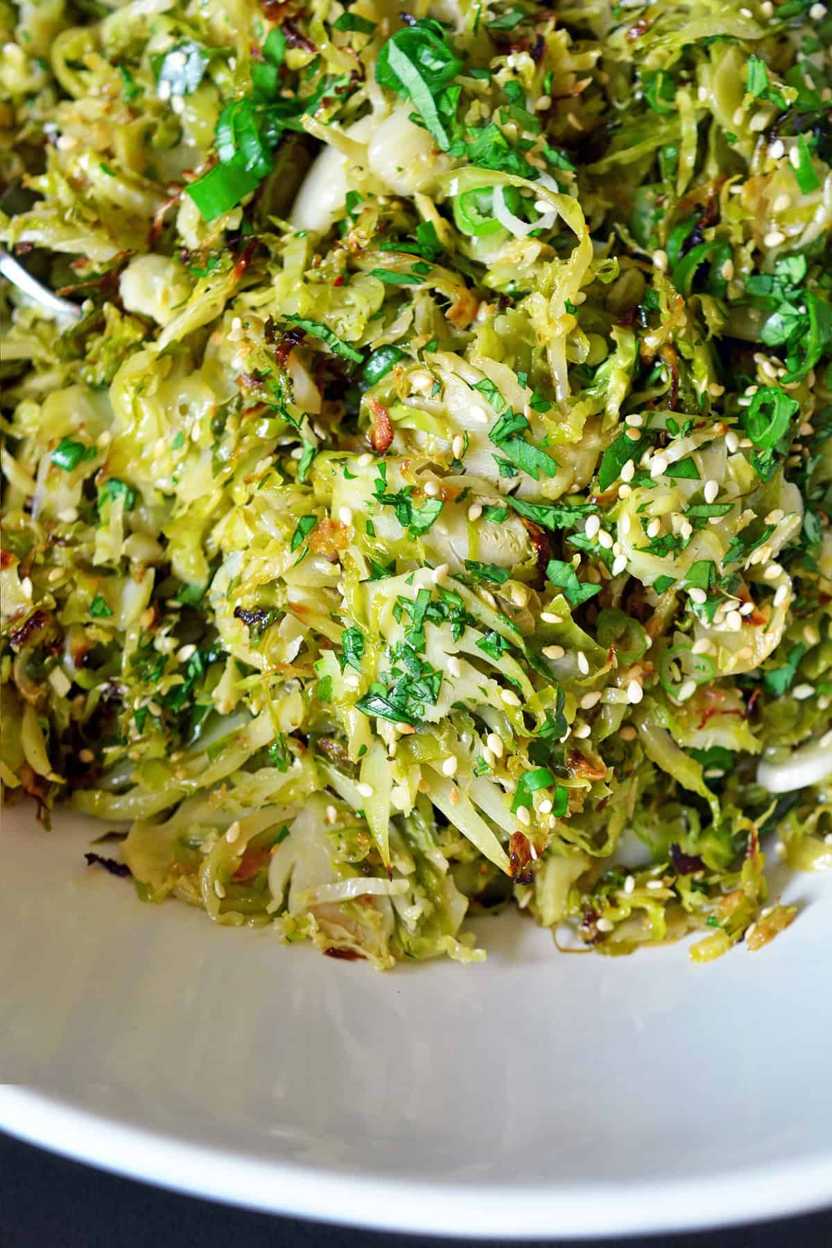 An overhead closeup shot of a white bowl filled with Roasted Brussels sprouts salad with Asian dressing topped with green onions and toasted sesame seeds.
