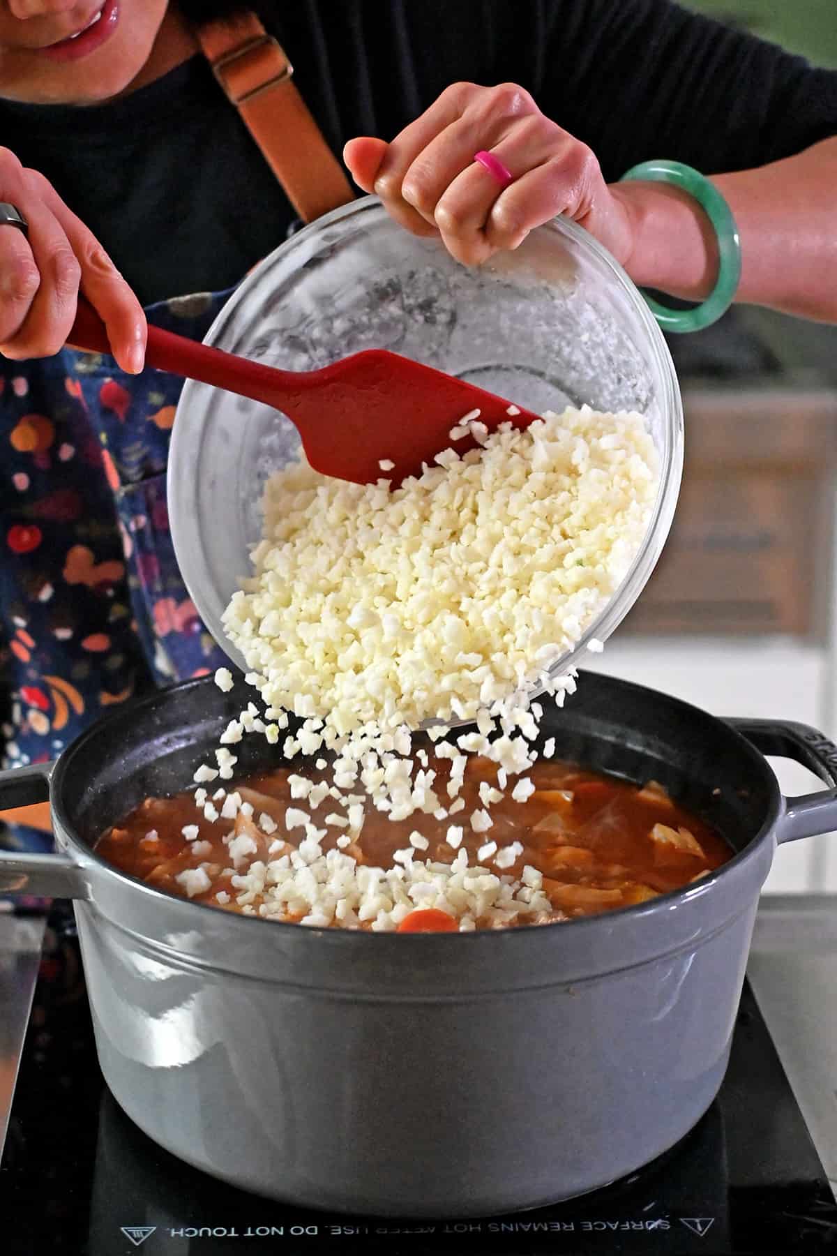 Adding a bowl of frozen riced cauliflower to a simmering pot of cabbage roll soup.