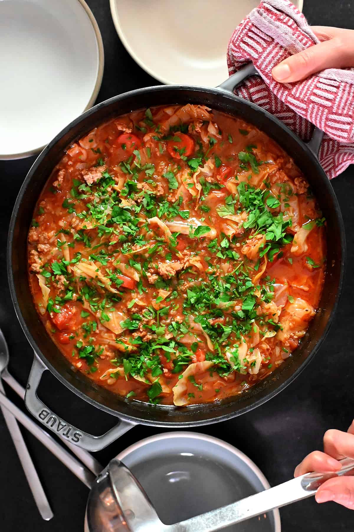 An overhead shot of a pot of cabbage roll soup topped with minced Italian parsley, next to some serving bowls and a ladle.