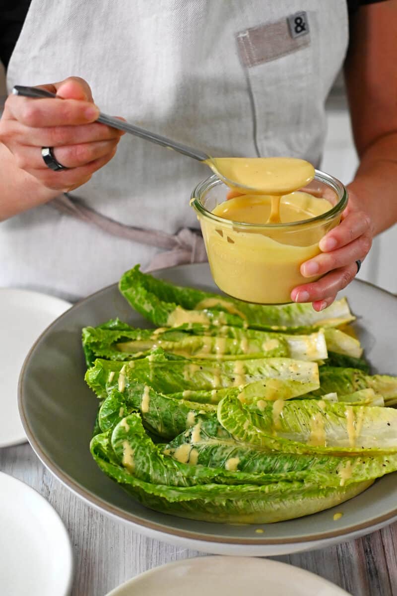 Someone adding dairy free caesar dressing to a shallow bowl filled with romaine lettuce leaves