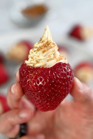 A hand is holding up a paleo and vegan cheesecake stuffed strawberry topped with gluten-free cookie crumbs.