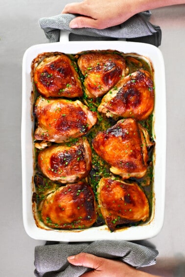 An overhead shot of two hands holding a white baking dish filled with golden brown chicken thighs and mushrooms.