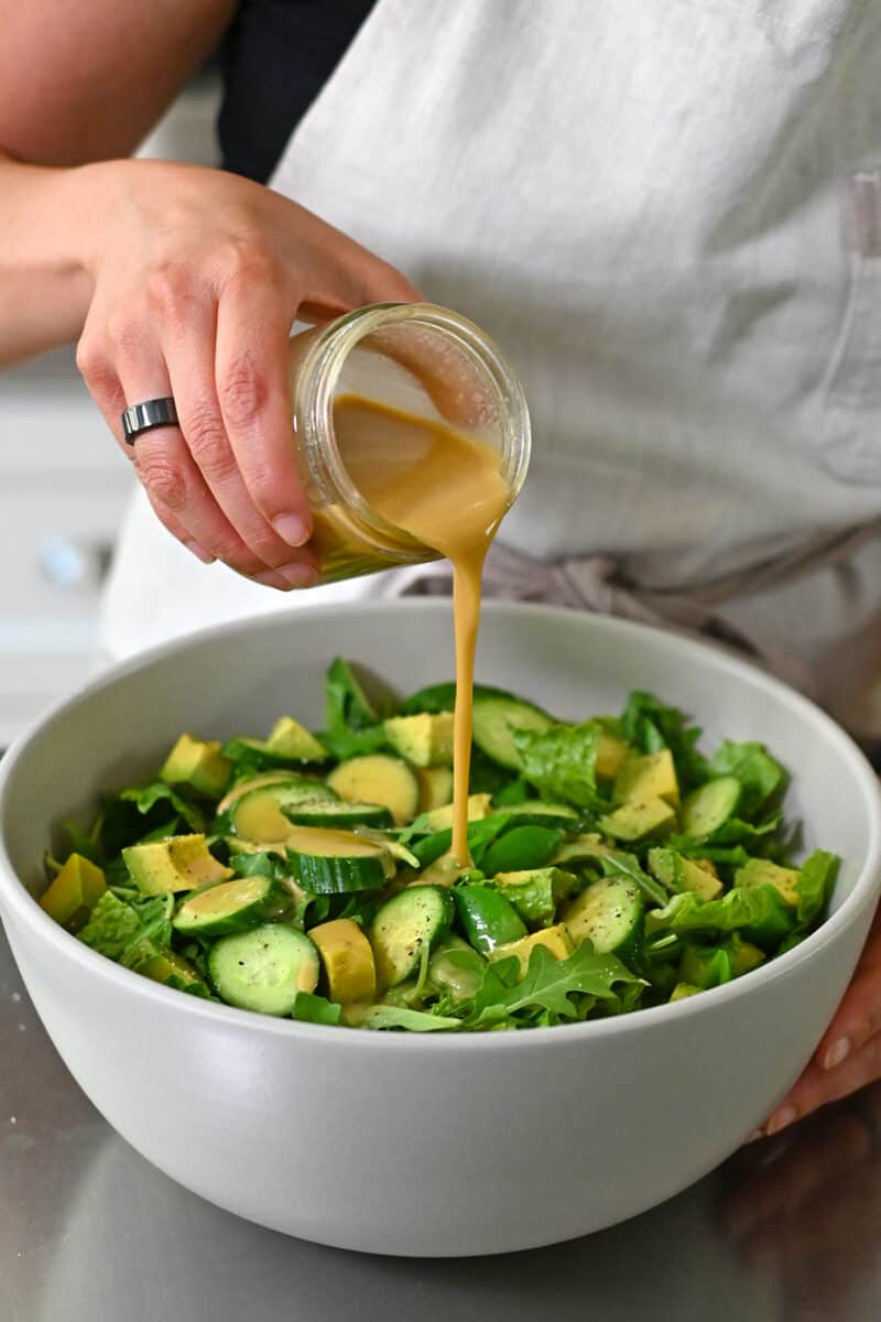 Pouring sesame ginger dressing into a large bowl of chopped salad.