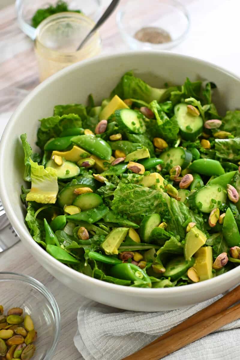 A closeup of a green chopped salad with salad greens, cubed avocado, sugar snap peas, sliced cucumbers, pistachios, and sesame ginger dressing.