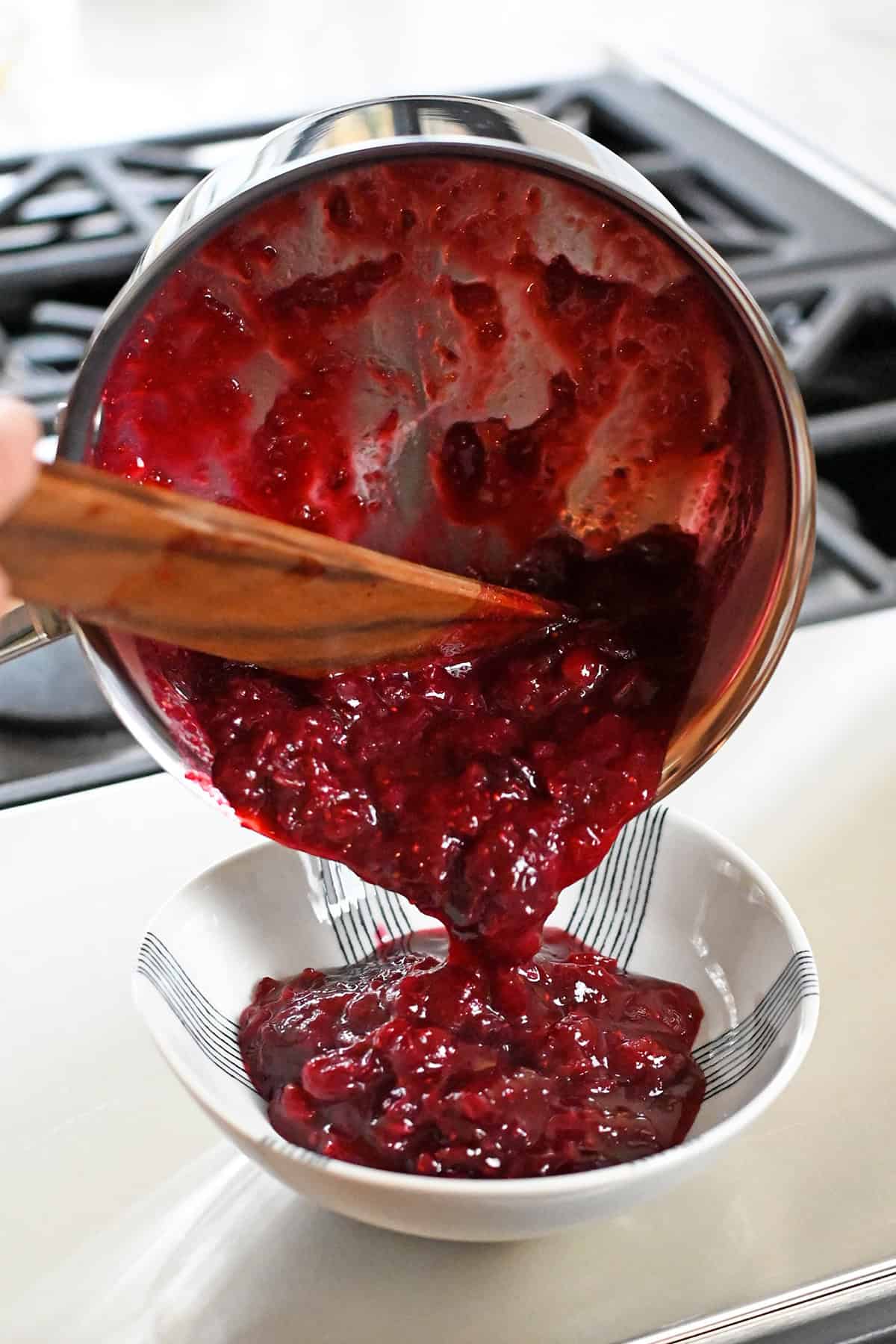 Pouring paleo cranberry sauce from a saucepan into a white serving bowl.