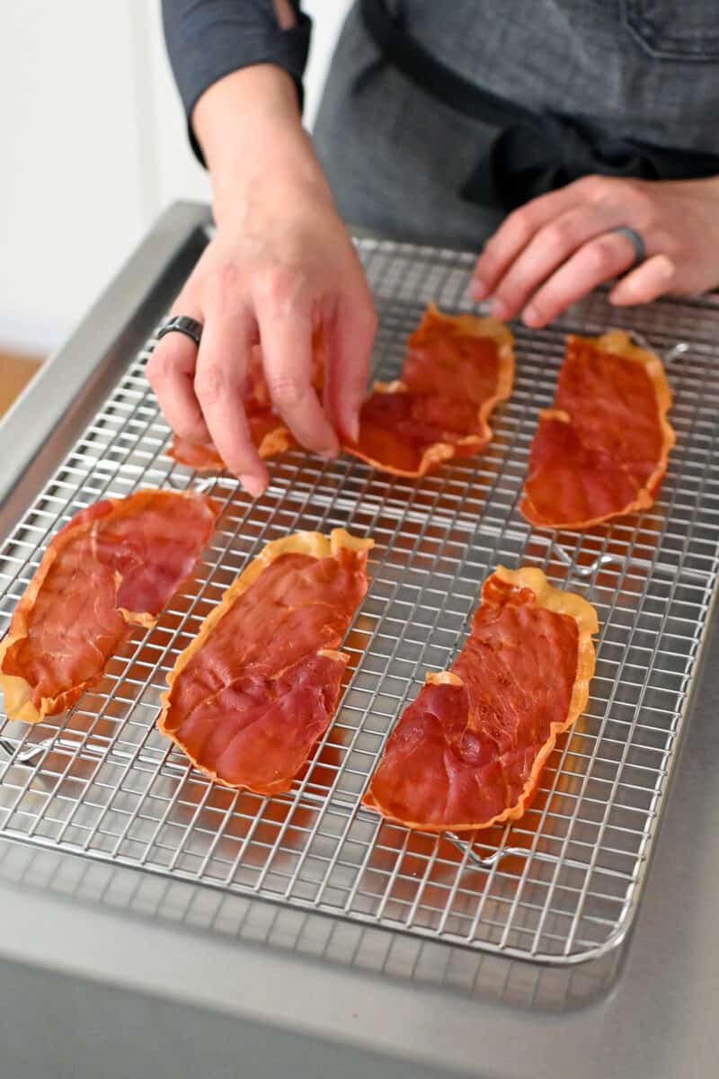 A hand is placing crispy prosciutto chips on a cooling rack