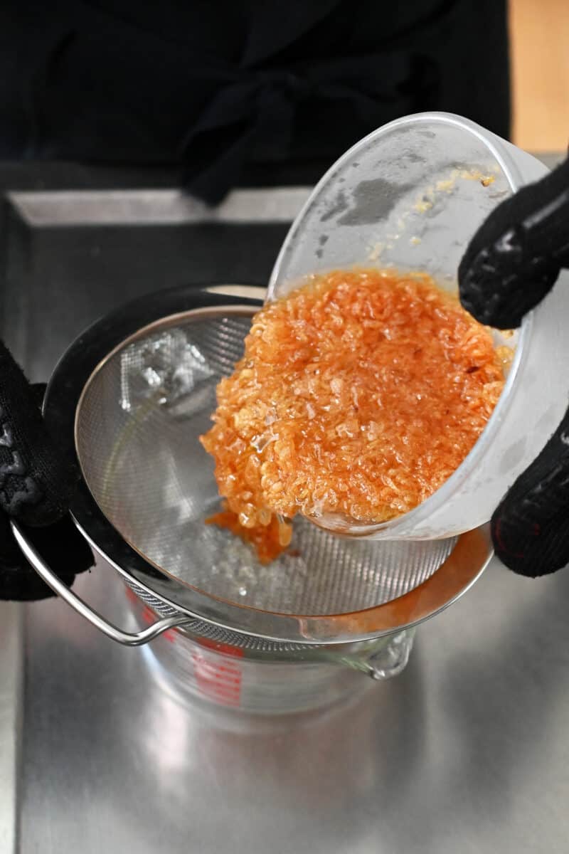 Pouring a bowl filled with crispy shallots and oil through a fine mesh sieve into a bowl.