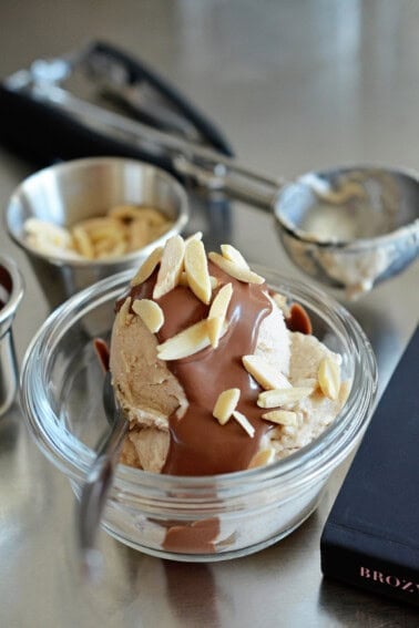 A side view of a scoop of homemade ice cream topped with chocolate ganache and almond slivers next to the topping ingredients, a dirty scooper, and a cookbook.