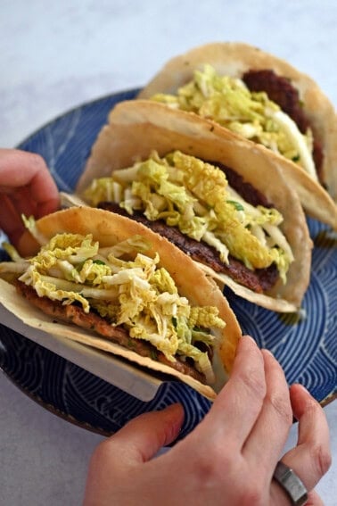 Two hands are picking up a dumpling smash taco from a blue plate filled with three of them.