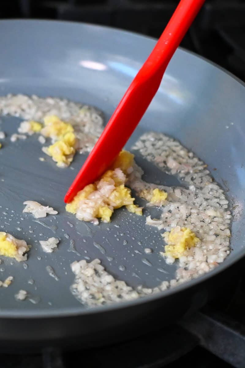 A closeup of a red silicone spatula pushing minced ginger and finely diced shallots in a large skillet.