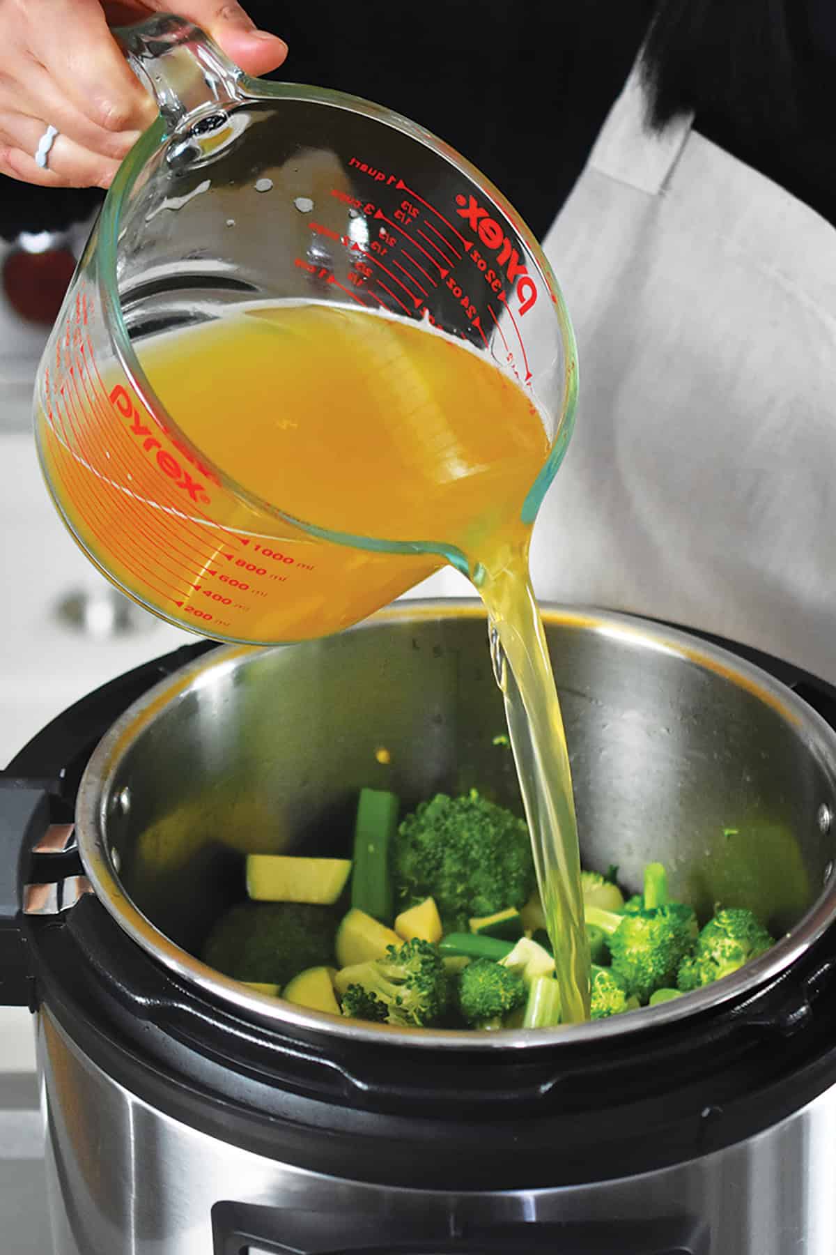 Pouring chicken broth into a pot with broccoli, green onions, zucchini, and apple to make green soup.