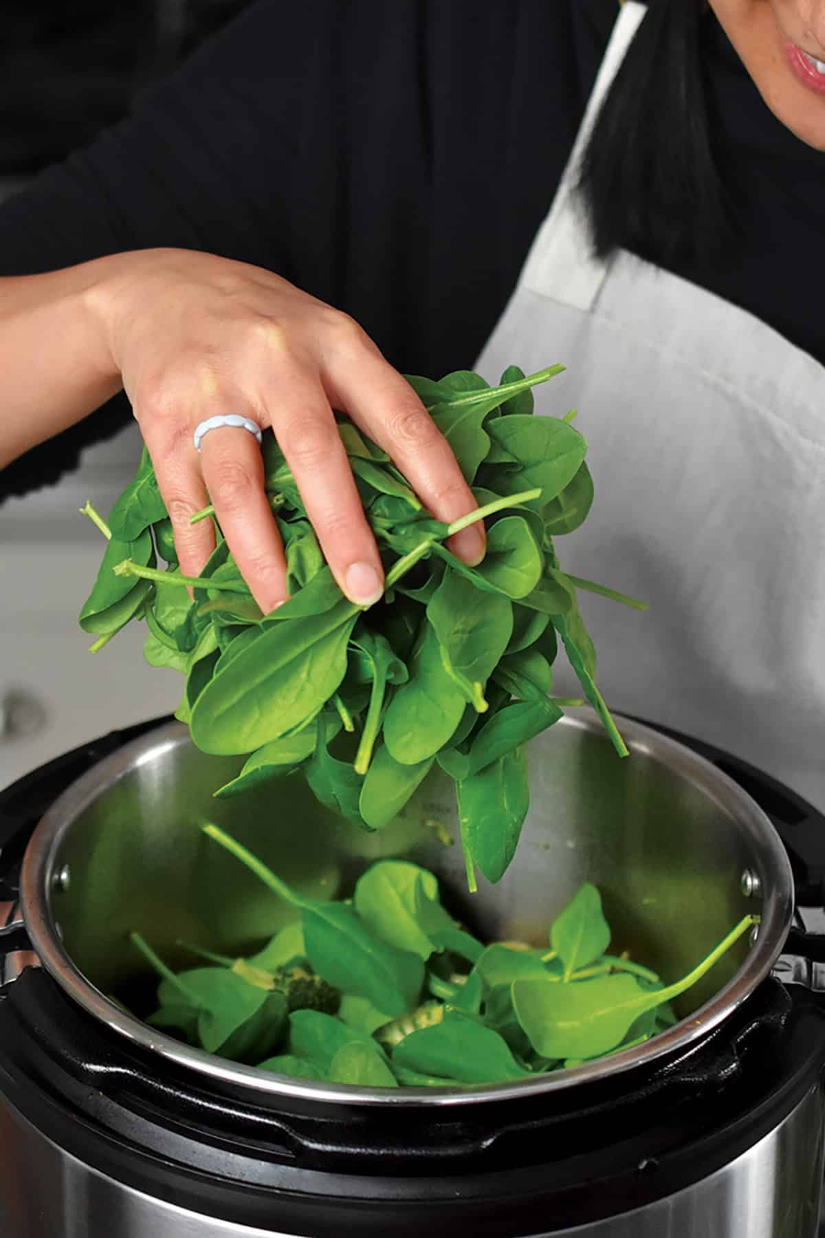 A hand is adding raw baby spinach to an open Instant Pot filled with the cooked green soup ingredients.