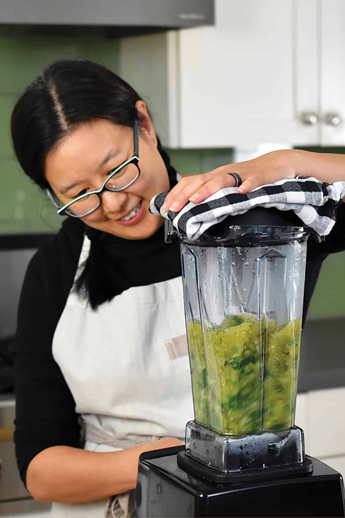 A smiling Asian woman is blending green soup in a Vitamix.