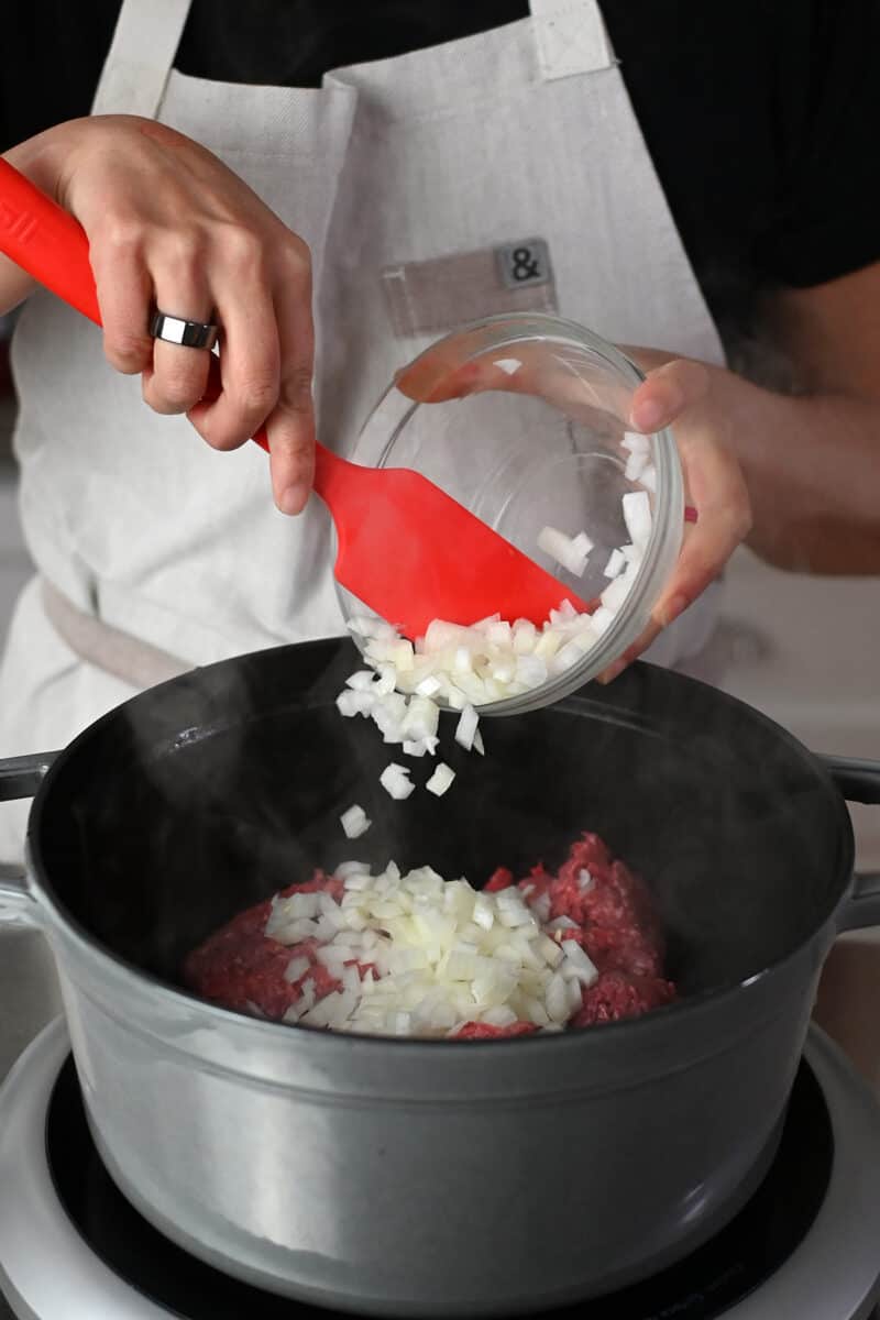 Adding diced onions to a gray stockpot filled with ground beef. 