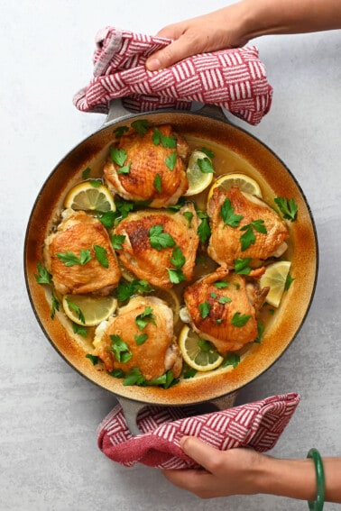 An overhead shot of two hands holding a skillet filled with lemon garlic chicken thighs with two red and white kitchen towels.