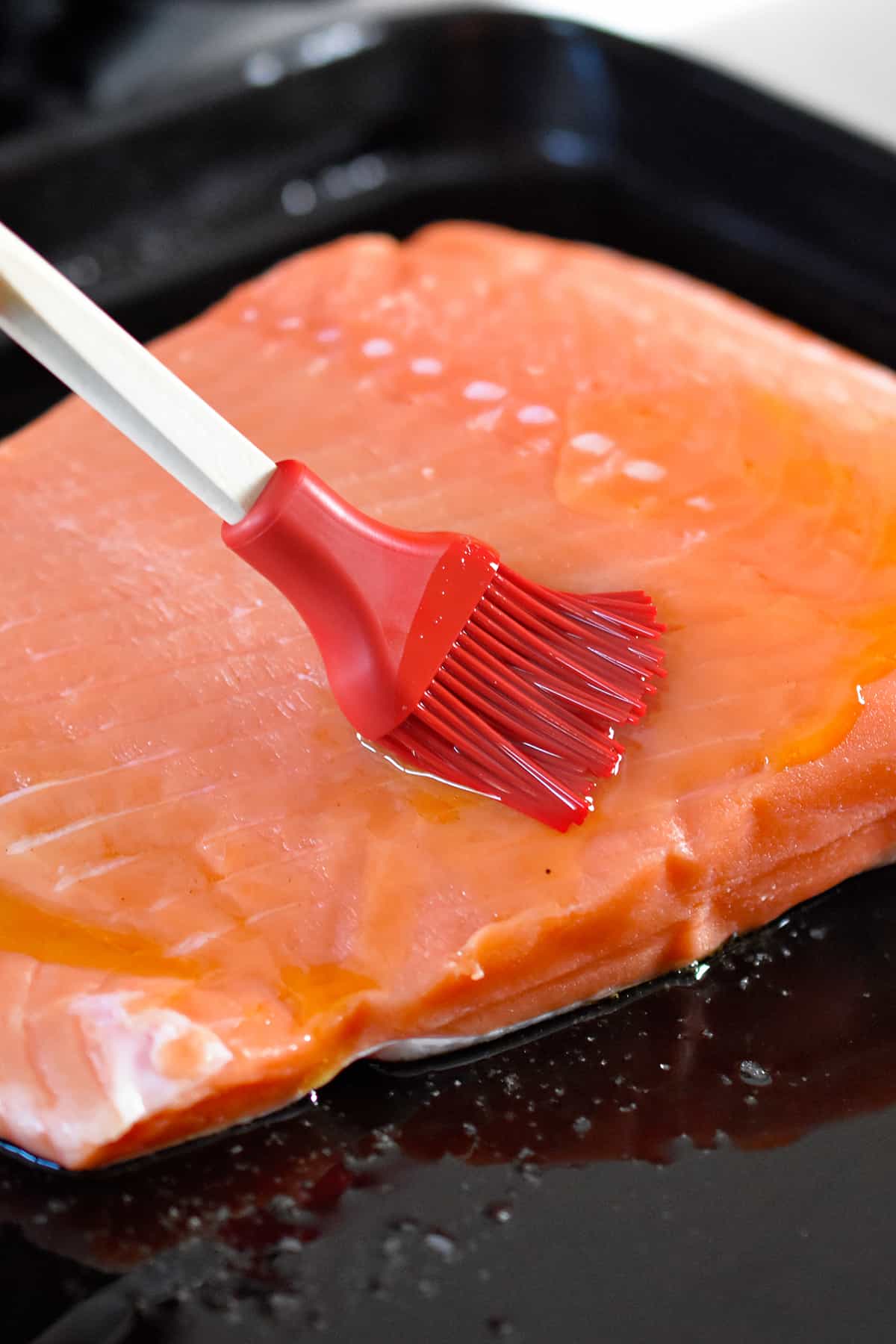 A red silicone brush is applying melted ghee on a raw salmon fillet on a black rimmed baking sheet.