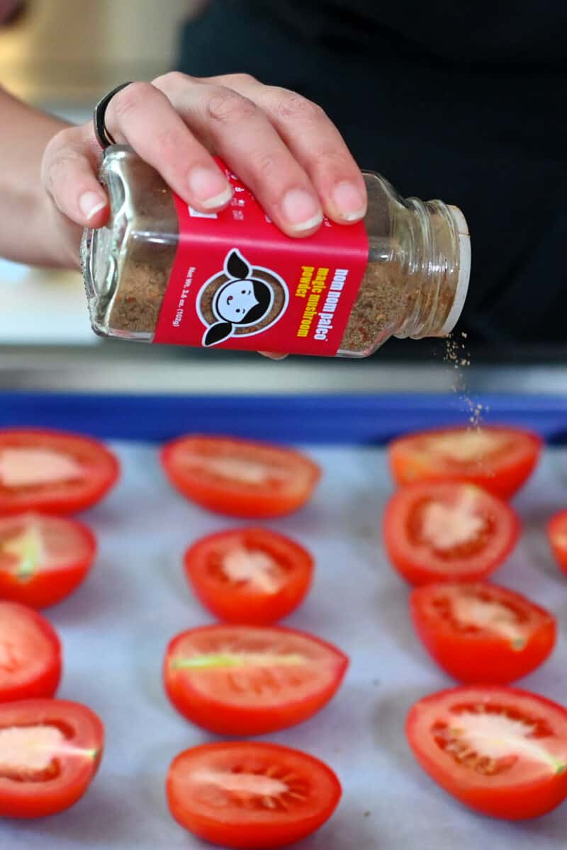 Sprinkling a jar of Magic Mushroom Powder onto halved plum tomatoes on a baking sheet.