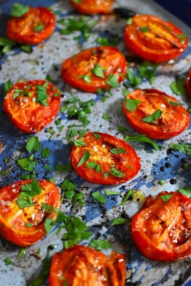 A closeup shot of oven roasted tomatoes on a blue baking sheet topped with chopped Italian parsley.