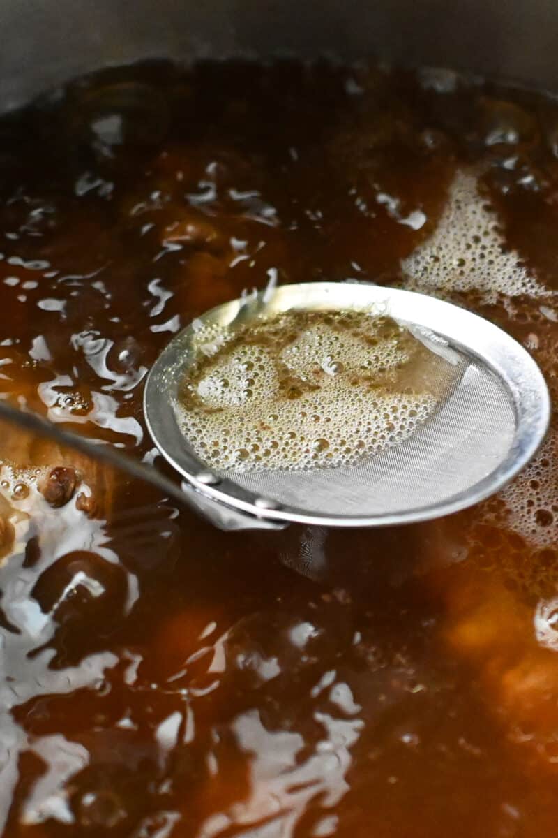 A small metal sieve is skimming the top of simmering oxtail broth and removing scum.
