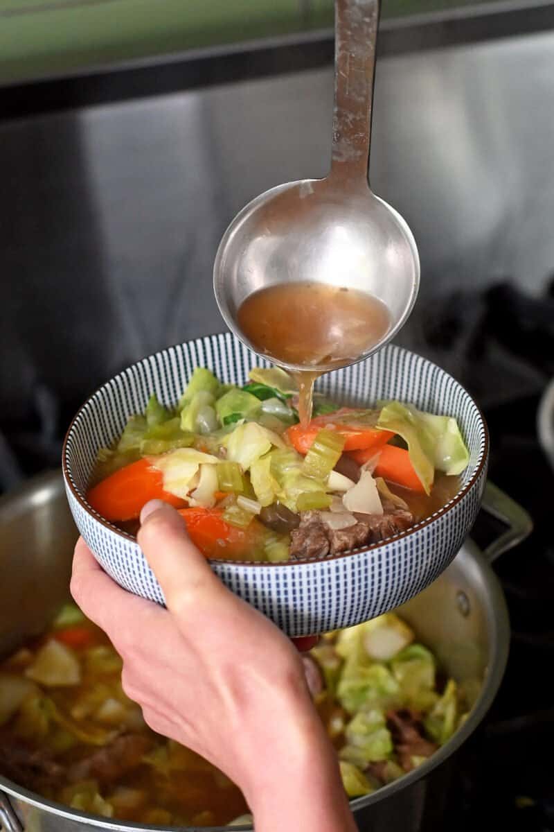 Ladling bowl of oxtail soup into a blue and white bowl.