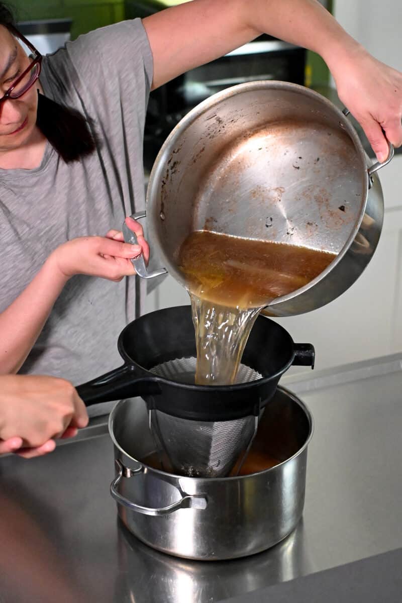 Straining oxtail soup through a sieve into a large pot.