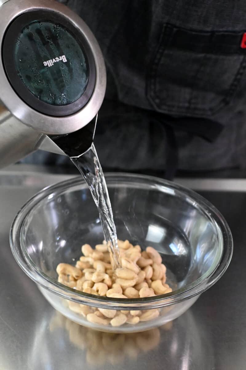 Hot water from a kettle is being poured on top of raw cashews in a clear glass bowl.