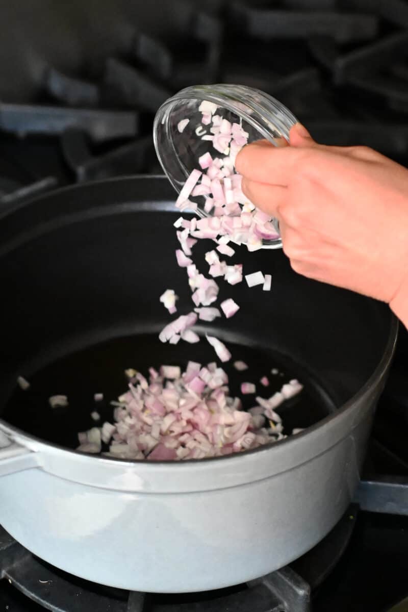 A bowl of chopped shallots is being poured into a gray dutch oven to make pea soup