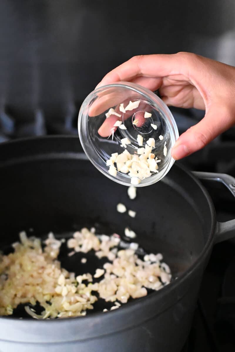 Adding chopped garlic to a gray stock pot with sautéed chopped shallots inside